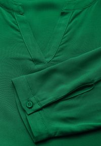 Blusa verde con scollatura a V e maniche lunghe. Il tessuto ha una texture liscia, con un dettaglio di bottone sul polsino e sulle cuciture.