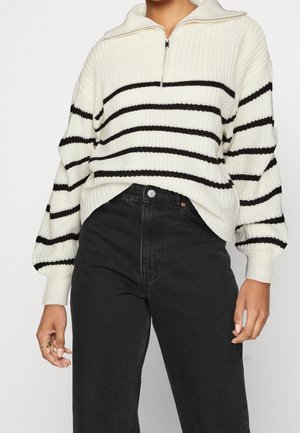 Femme portant un pull en maille blanc à rayures noires horizontales et une fermeture éclair sur le devant, associé à un pantalon noir taille haute.