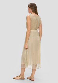 Robe plissée beige avec une texture brillante, sans manches, et une ceinture à la taille. Associée à des sandales plates dorées, vue de dos.