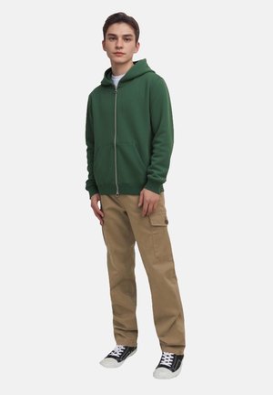 Green zip-up hoodie with a front pocket, paired with beige cargo pants and black sneakers featuring white soles. Simple design, casual fit.