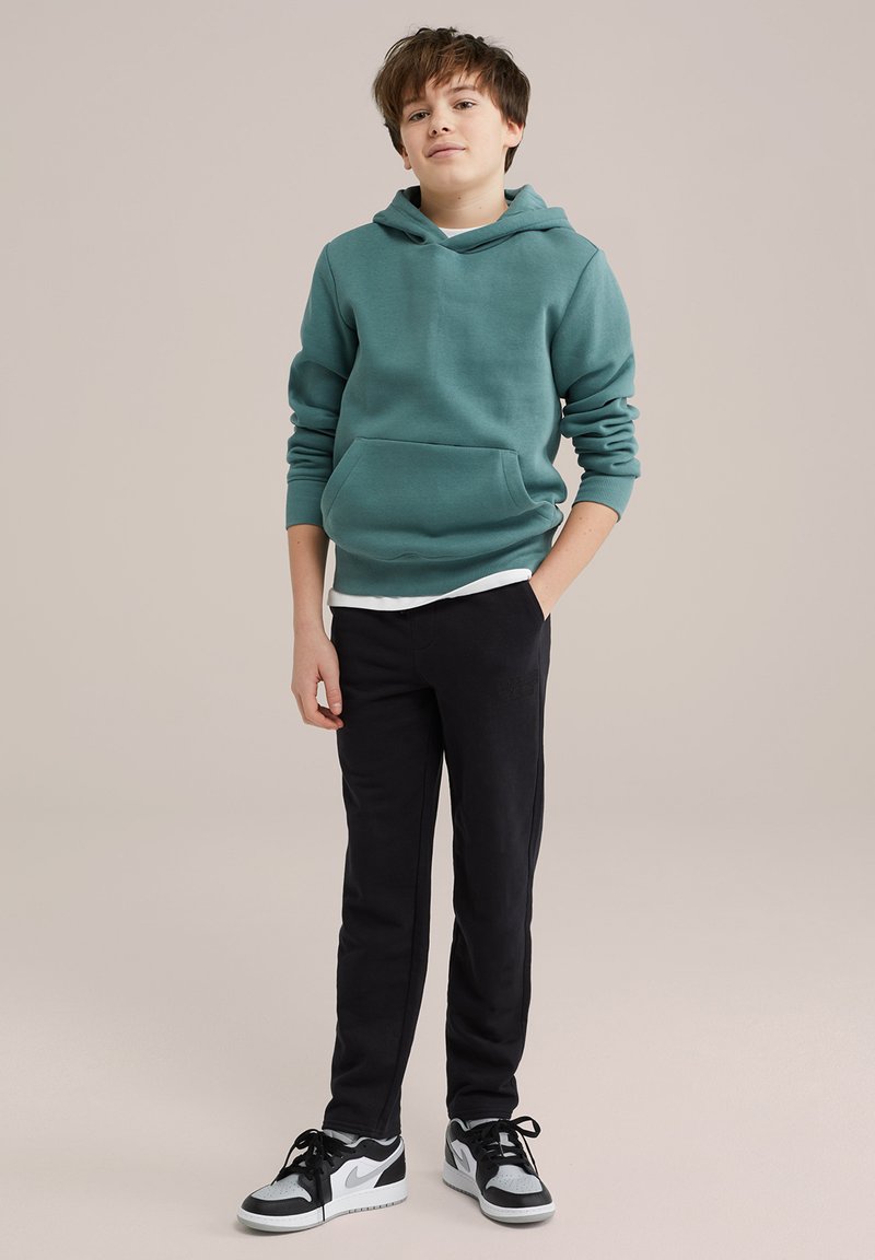 Tiržna zelena kapuca s kengurskim žepom, složena preko bele majice; v kombinaciji s črnimi hlačami in črno-belimi supergami.