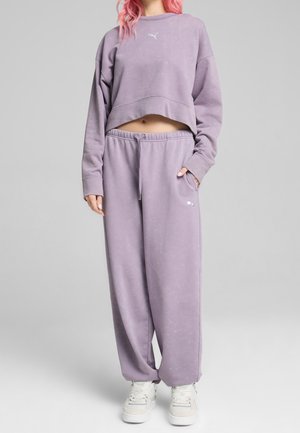 Personne ayant les cheveux roses portant un sweat-shirt court lavande et un pantalon de survêtement large assorti avec des baskets blanches.