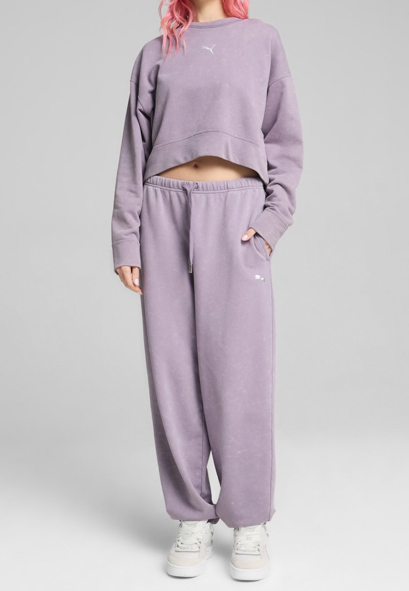 Personne ayant les cheveux roses portant un sweat-shirt court lavande et un pantalon de survêtement large assorti avec des baskets blanches.