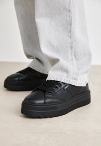 Mustat nahkakengät, joissa paksut pohjat, pyöreä kärki, tasaiset nauhat ja merkitty sivuelementti. Yhdistetty vaaleanharmaisiin kuluneisiin farkkuihin.