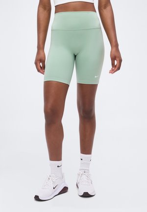 Athlète portant un short ajusté vert clair, des chaussettes blanches Nike et des baskets blanches Nike, debout devant un fond clair uni.