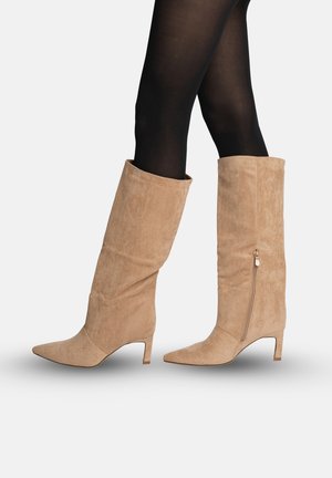 Beige suède kniehoge laarzen met zijrits, gedragen over zwarte maillots, gezien van opzij met lage kittenhakken en puntige neuzen.