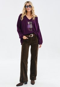 Fioletowy sweter z dekoltem w serek z grafiką dwóch kotów, w połączeniu z brązowymi szerokimi spodniami z kordurowej tkaniny i brązowymi botkami na obcasie.