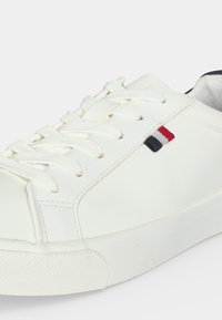 Λευκό χαμηλό sneaker με λευκά κορδόνια, υφασμάτινη σόλα με υφή και μικρή υφασμάτινη ετικέτα σε κόκκινο, λευκό και μπλε στην πλευρά.