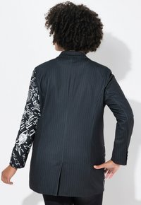 Blazer nero con righe sottili, dotato di maniche con motivo contrastante. Presenta una ventola sul retro e una vestibilità sartoriale per un indossare strutturato.
