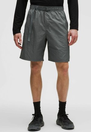 Mężczyzna noszący ciemnoszare casualowe szorty z regulowanym paskiem, czarne buty sportowe i czarne skarpetki, stojący na jednolitym tle.