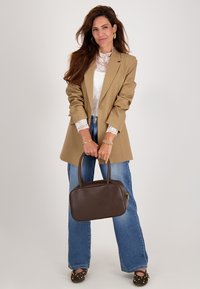 Bruine leren handtas met dubbele handvaten, rechthoekige vorm, gladde textuur, gouden ritsdetails. Model draagt een beige blazer en blauwe jeans.