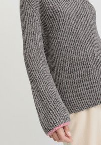 Grau gestrickter Pullover mit gerippter Textur, der weit geschnittene Ärmel und einen hellrosa Akzent an den Bündchen aufweist.