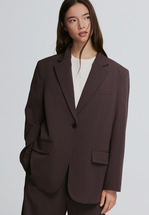 Mladá žena s dlhými tmavými vlasmi, oblečená v nadrozmernom tmavom pruhovanom blejzri a zodpovedajúcich nohaviciach cez bielu košeľu, hľadí do strany.