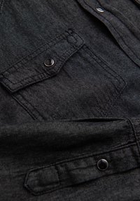 Chemise en jean noire avec une poche à rabat à fermeture pression, tissu texturé et détails de couture visibles. Matière lisse et durable.