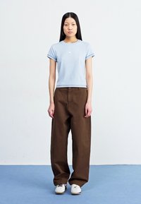 T-shirt azul-claro de manga curta com um pequeno logotipo, combinado com calças largas castanhas e ténis brancos. Design simples, ajuste descontraído.