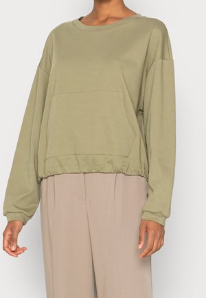 Persoon draagt loszittende olijfgroene lange mouwen sweatshirt met voorzak en beige broek met wijde pijpen tegen een effen achtergrond.
