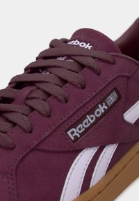 Sapatilhas de camurça bordô com detalhes brancos, atacadores texturizados e uma sola de borracha castanha. Apresenta o logótipo da Reebok, realçando elementos de design retro.