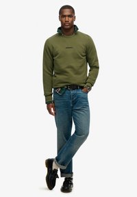 Olijfgroene sweatshirt met geborduurd logo, gelaagd over een geruite shirt, gecombineerd met blauwe jeans en zwarte schoenen met een gele zool.