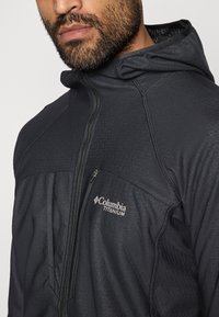 Fekete kapucnis dzseki cipzárral az elülső részen, texturált anyaggal, a Columbia Titanium logóval a bal oldalon. Sleek, modern dizájnja van.