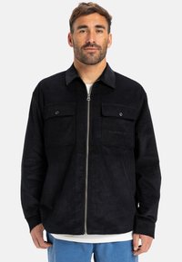 Giacca in corduroy nera con zip frontale, due tasche sul petto con bottoni e dettagli del logo discreti. Presenta un colletto e una vestibilità rilassata.