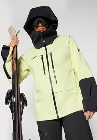 Veste de ski jaune vif et noire avec un col haut, des poches zippées et une capuche réglable. Modèle tenant des skis, portant de grandes lunettes de ski.