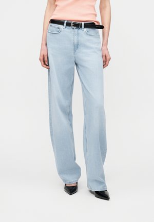 Person iført lyseblå bredbenede jeans, sort bælte, ferskenfarvet top og sorte spidse sko mod en ensfarvet hvid baggrund.