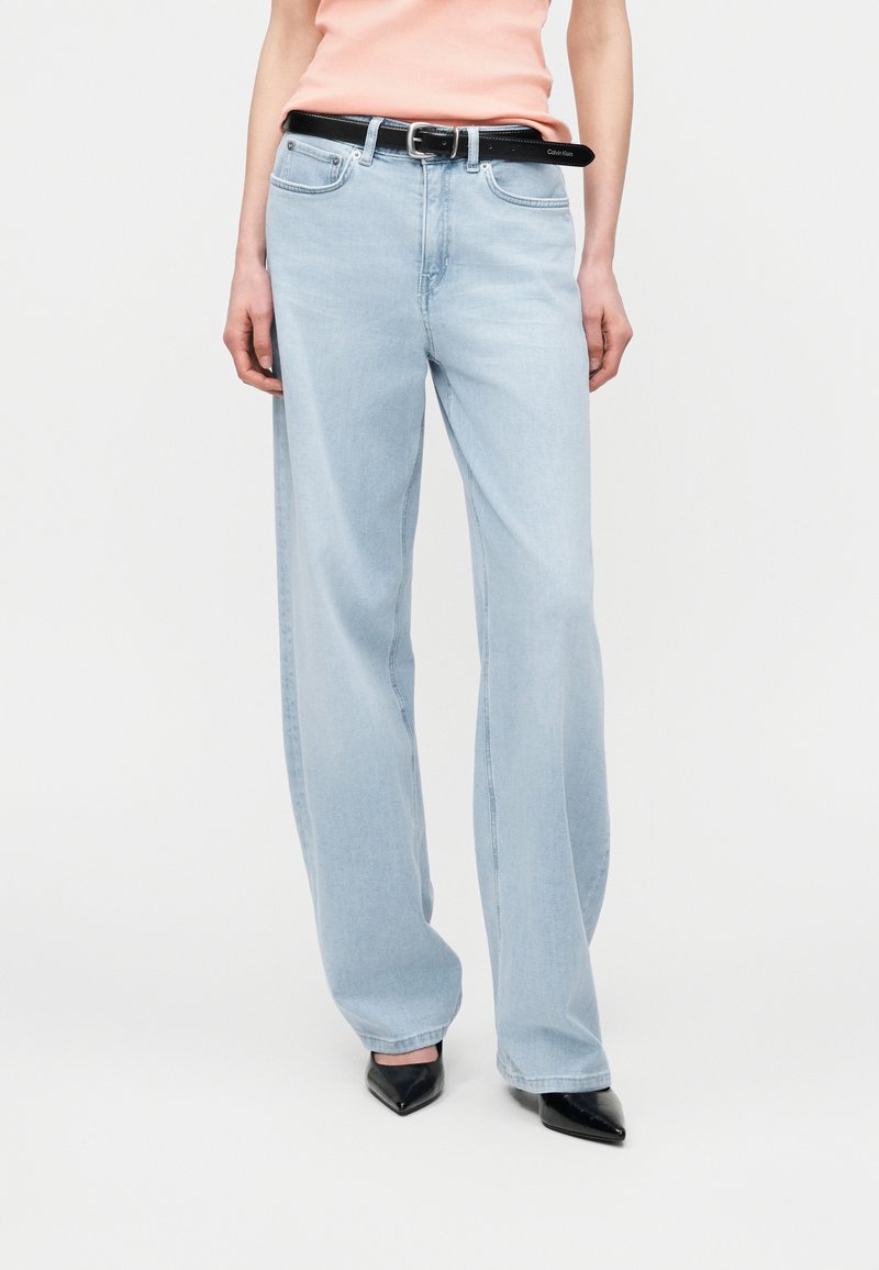 Persona vistiendo jeans acampanados azul claro, cinturón negro, blusa melocotón y zapatos negros de punta afilada, sobre un fondo blanco liso.