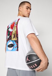 Biela bavlnená tričko s grafickou potlačou na chrbte s leopardským motívom, basketbalmi a logom Nike. Model drží textúrovaný basketbal.
