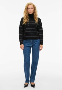 Črn pulover s turtleneck ovratnikom in sivimi vodoravnimi črtami, v kombinaciji z modrimi naravnost krojenimi kavbojkami in črnimi špicastimi petami. Gladka tekstura in oprijet dizajn.