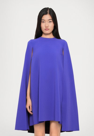 Frau mit langen schwarzen Haaren, die ein ärmelloses lebhaft lilafarbenes Kapuzenkleid trägt und vor einem weißen Hintergrund steht.