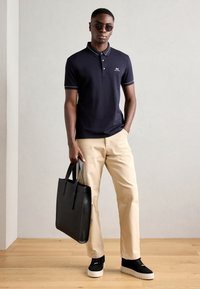 Marineblauwe polo met witte accenten, beige broek, zwarte totebag met handvatten en zwarte sneakers met een witte zool. Soepele stof en casual pasvorm.