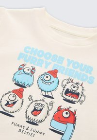 Cremefarbener T-Shirt mit Cartoon-Monstern in verschiedenen Farben und verspielten Ausdrücken. Der Text lautet "Wähle deine pelzigen Freunde."