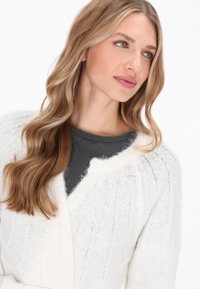 Fehér, bordázott kötött kardigán, puha textúrával és nyitott elülső résszel, sötétszürke, testhezálló felsőre rétegezve. Hosszú, hullámos haj és természetes smink.