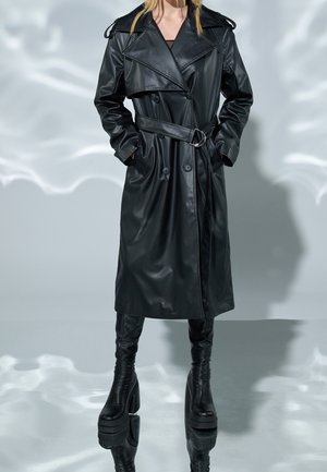 Personne portant un trench-coat en cuir noir ceinturé avec des poches latérales et des bottes à talons compensés noires, debout sur un sol réfléchissant.