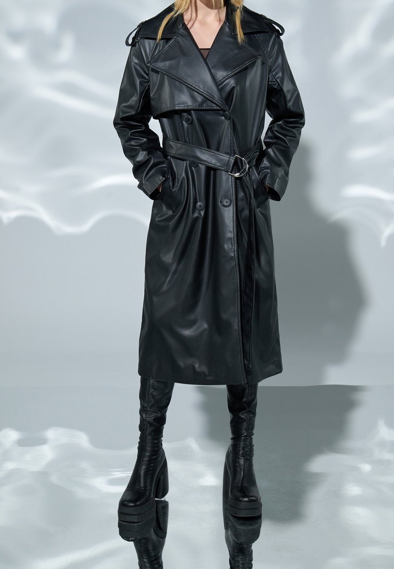 Personne portant un trench-coat en cuir noir ceinturé avec des poches latérales et des bottes à talons compensés noires, debout sur un sol réfléchissant.