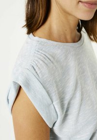 Camisa de manga corta azul claro con puños enrollados, que presenta sutiles detalles de pliegues y una tela suave y texturada con un ligero brillo.