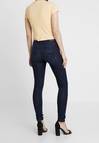 Kvinna i mörkblå slim fit-jeans, beige kortärmad topp och svarta högklackade sandaler, som står mot en enfärgad vit bakgrund.