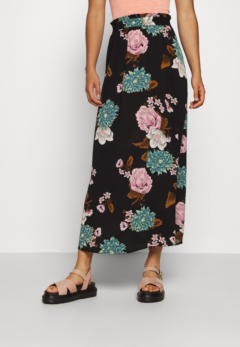 Femme portant une jupe midi noire à fleurs roses et bleues, un haut sans manches pêche, et des sandales compensées roses.