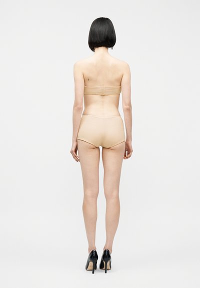 Femme aux cheveux noirs courts portant un soutien-gorge beige sans bretelles et un slip assorti, avec des talons hauts noirs, debout de dos sur fond blanc.