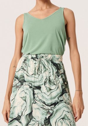 Femme portant un haut sans manches vert clair associé à une jupe taille haute avec un motif marbré vert et blanc.