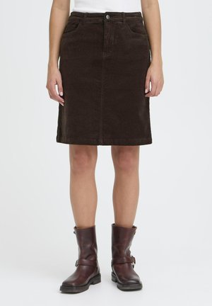 Mujer con una falda de pana marrón oscuro hasta la rodilla y botas de cuero marrón oscuro hasta la mitad de la pantorrilla, de pie frente a un fondo liso.