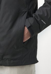 Sort vandtæt jakke med et tekstureret stof, justerbare manchetter med Velcro og en sidelomme med lynlås.