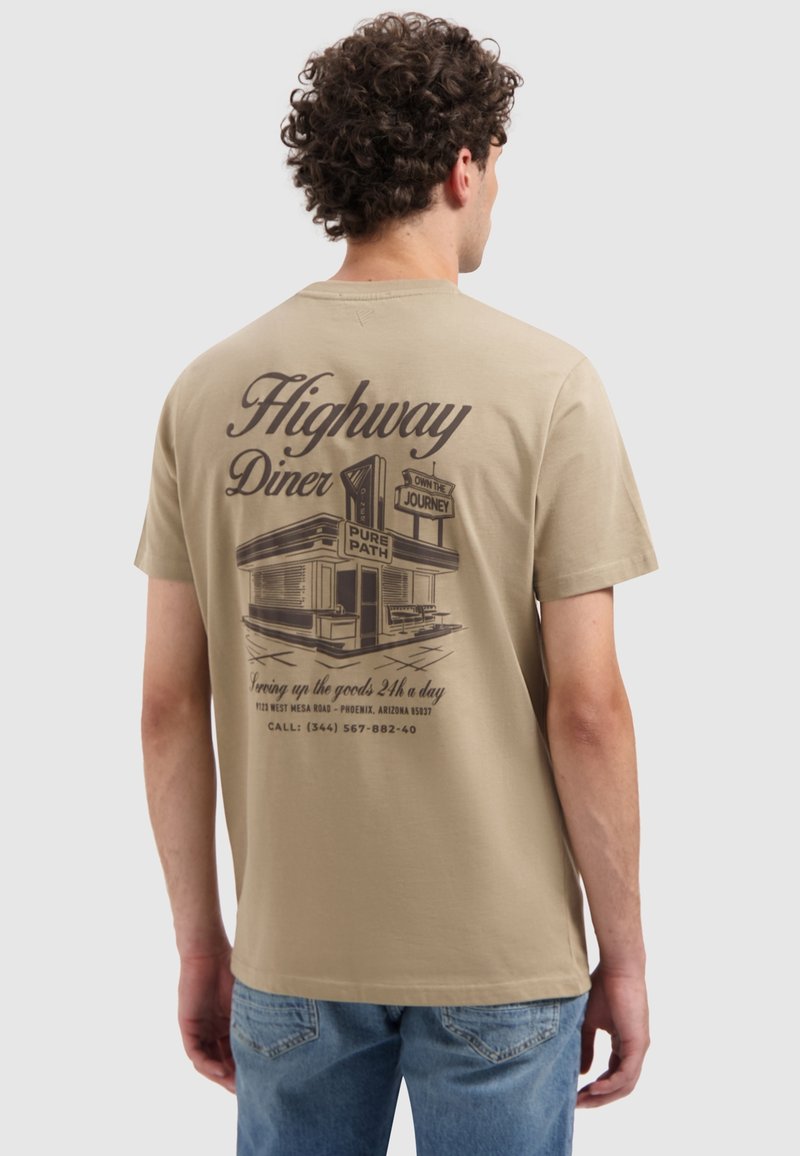 Man met een beige T-shirt met een vintage diner grafiek en tekst "Highway Diner" en contactgegevens op de achterkant, blauwe spijkerbroek.
