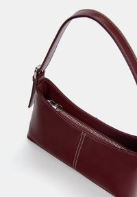 Burgunderfarbene Lederhandtasche mit schmalem Design, ausgestattet mit einem Reißverschlussfach, weißem Steppnähten und einem abnehmbaren Schultergurt.