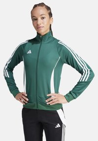 Giacca verde con zip realizzata in tessuto liscio, con strisce bianche sui lati e un logo ricamato sul petto. Vestibilità comoda per l'abbigliamento sportivo.