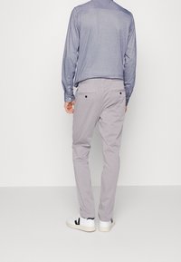 Uomo che indossa pantaloni grigi chiari, camicia a maniche lunghe blu e sneakers bianche, in piedi di fronte a un muro bianco, visto da dietro.