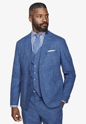 Mężczyzna z brodą ubrany w niebieski, fakturowany garnitur trzyczęściowy, koszulę w paski i krawat w wzory, patrzący w prawo, z ręką w kieszeni.