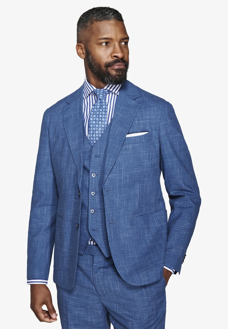 Uomo con barba che indossa un completo blu a tre pezzi dal tessuto strutturato, camicia a righe e cravatta fantasia, guarda verso destra con la mano in tasca.