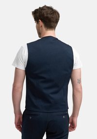 Chaleco marino con un diseño ajustado, hecho de tejido texturizado, cuenta con una parte trasera lisa y detalles de costura sutiles. Se lleva sobre una camiseta blanca.