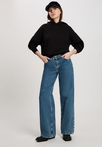 Czarna włóczkowa bluza z kapturem i szerokie niebieskie dżinsy. Tekstura materiału jest gładka, z subtelnym detalem logo na przodzie. Czarne obuwie dopełnia stylizację.